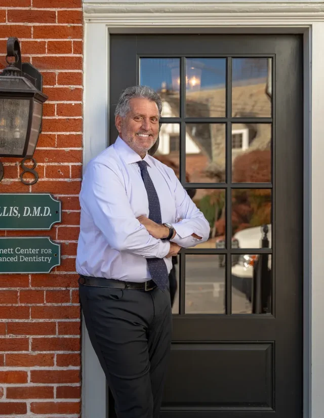 Dentist in a white shirt and tie leaning against his office door beside a brick wall