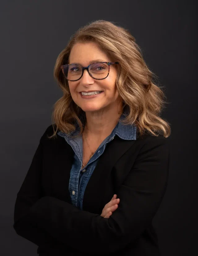 Woman in glasses and a black blazer, arms crossed, studio headshot on charcoal