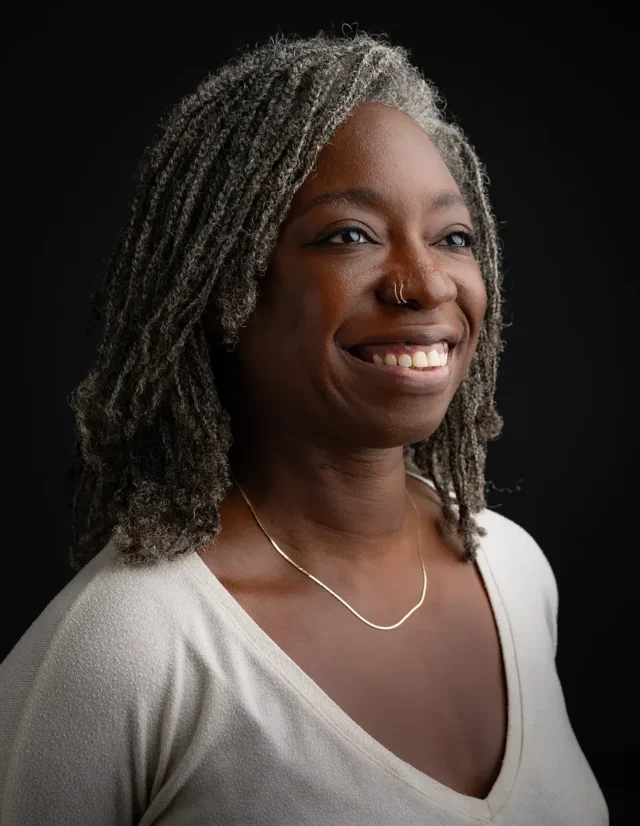 Smiling woman with gray locs in a cream top on a black background