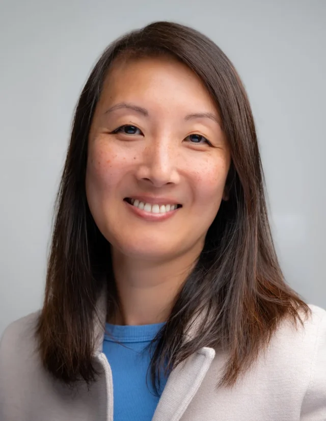 Smiling woman in a light jacket and blue top, headshot on soft gray